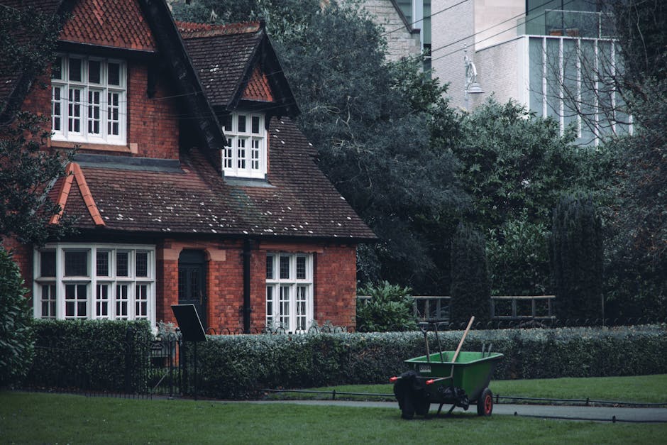 A traditional red-brick house with a sloped tiled roof and white-framed windows set amidst a well-maintained garden. The garden features neatly trimmed hedges, a lush green lawn, and various mature tr